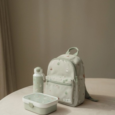 Mochila, garrafa e lancheira infantil verdes claros com padrões infantis sobre uma mesa clara.