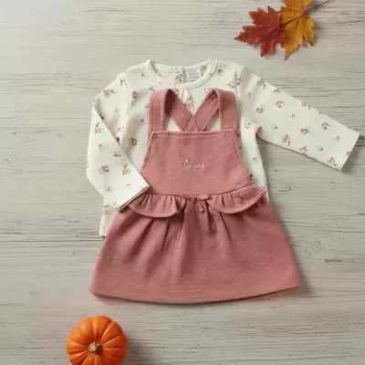 Conjunto infantil com vestido rosa e camisa branca floral sobre superfície de madeira clara com folhas de outono e abóbora