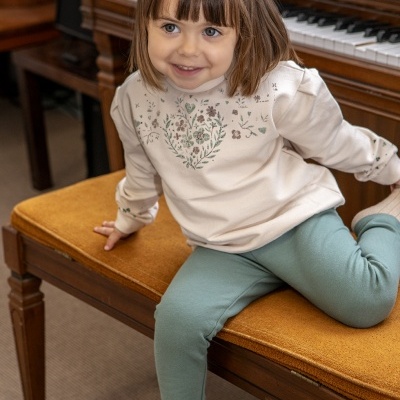 Menina sentada no banco acolchoado laranja, com camisola creme estampada e calças verdes perto de piano de madeira.