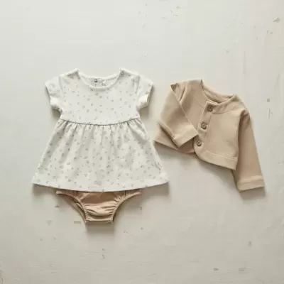 Conjunto de bebé com vestido branco, fralda bege e casaco bege.