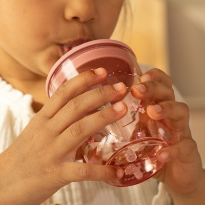 Copo infantil plástico com tampa e palhinha rosa decorado com flores, segurado por criança para beber.