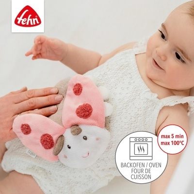 Bebé com vestido branco segurando brinquedo macio rosa e bege em forma de borboleta com manchas vermelhas e etiqueta Fehn.