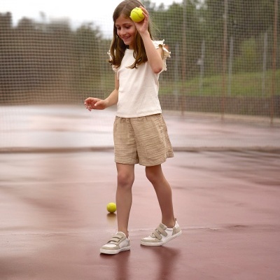 Rapaz de ténis bege e branco com velcro, calções e t-shirt branca com folhos segurando bola de ténis amarela num campo de ténis