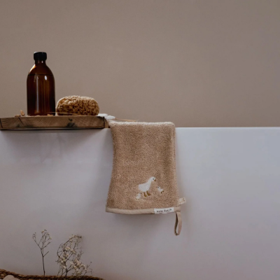 Toalha de mão bege com bordado de pato pendurada numa banheira branca com suporte de madeira e frasco âmbar