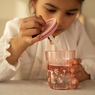 Copo infantil rosa com desenhos de veado e flores, com tampa e palhinha transparentes, na mão de uma criança com blusa branca.