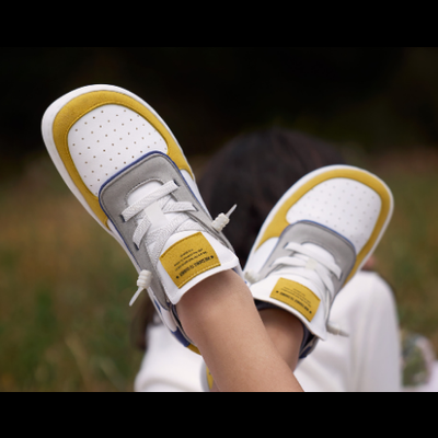 Ténis infantis nas cores branco, amarelo e cinzento com etiqueta amarela e atacadores brancos ao ar livre