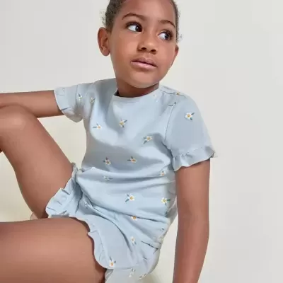 Criança vestindo conjunto azul claro com padrão de margaridas e folhos nas mangas e calções