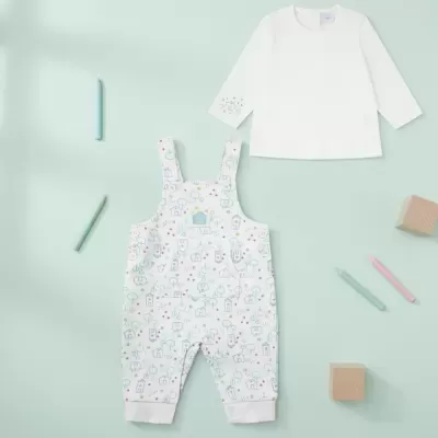 Conjunto de roupa infantil com camisa branca e macacão estampado, disposto sobre fundo verde claro