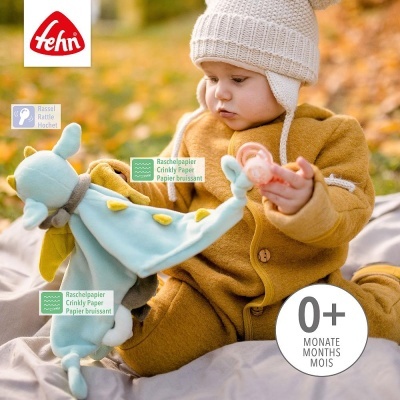 Bebé com uniforme castanho e gorro branco segurando brinquedo peluche azul no exterior com folhas amarelas