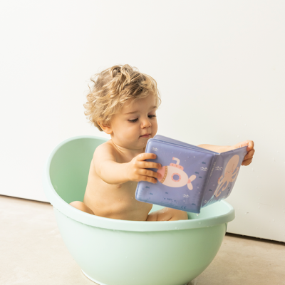 Bebé numa banheira verde menta a ler um livro de banho azul com desenhos marinhos.