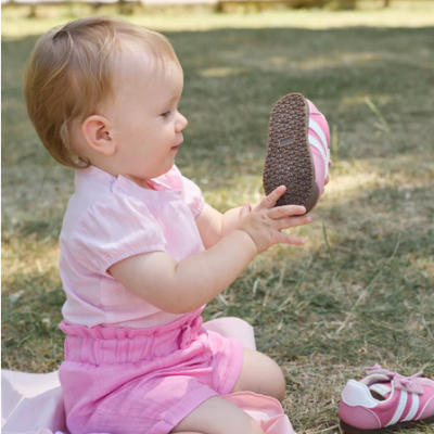 Criança sentada na relva com roupa rosa segurando sapato desportivo cor-de-rosa e branco