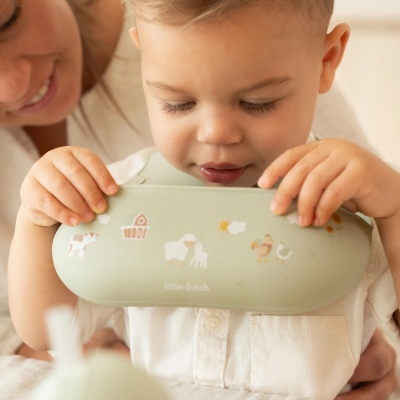 Criança a segurar objeto verde claro decorado com animais e texto, com adulto sorridente ao fundo