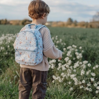 Criança com mochila azul claro com padrão de raposas e plantas, em campo com flores brancas.
