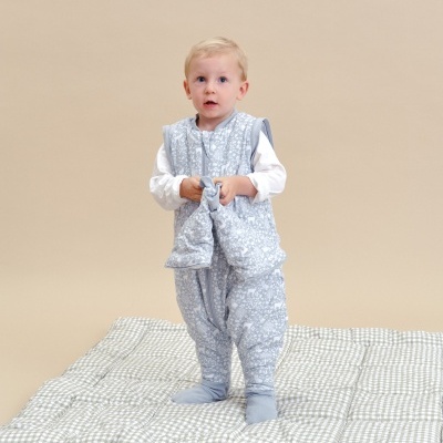 Criança vestindo saco de dormir azul claro com padrões brancos, em cima de tapete acolchoado xadrez verde e branco