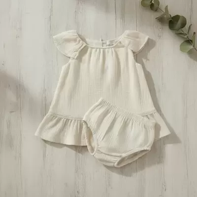 Conjunto de bebé creme com vestido leve e cuecas, textura riscada, sobre chão de madeira clara com ramo verde