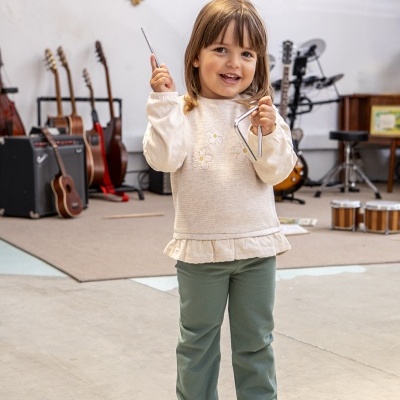 Criança com roupa creme e verde segurando triângulo musical em sala com instrumentos