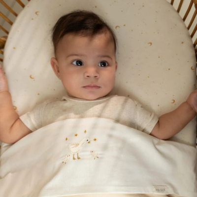 Bebé deitado em berço com pijama creme e manta branca bordada com patos e estrelas amarelas.