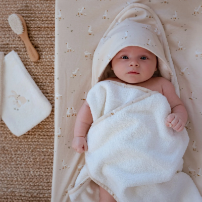 Bebé envolto em toalha de banho branca com capuz e padrão de patos, ao lado pente e toalha pequenas