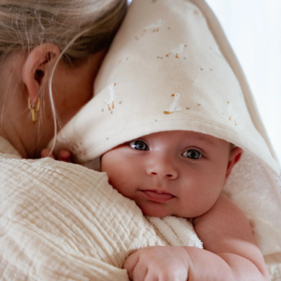 Bebé enrolado numa toalha com capuz creme e desenhos de patinhos, segurado por mulher com brinco dourado.