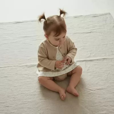 Bebé sentado num tapete bege com vestido branco e casaco bege