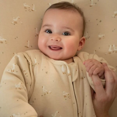 Bebé sorridente com pijama bege estampado com patinhos segurando a mão de um adulto