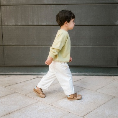 Criança vestida com casaco amarelo claro, calças brancas e sapatilhas castanhas a caminhar em chão de pedra, fundo cinzento.