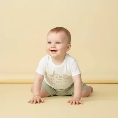 Bebé a sorrir vestido com t-shirt branca e calções verdes sentado em fundo amarelo