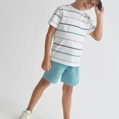 Menino com camisola riscada, calções azuis, boné e ténis brancos num fundo neutro