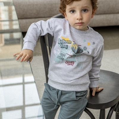 Criança com sweatshirt cinza estampado e calças cinza esverdeadas sentado numa cadeira preta junto a uma mesa de vidro