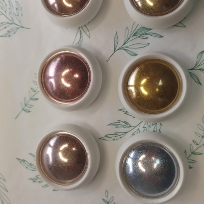 Seis cápsulas metálicas coloridas para cafeteira com base branca sobre superfície clara decorada com folhas verdes.