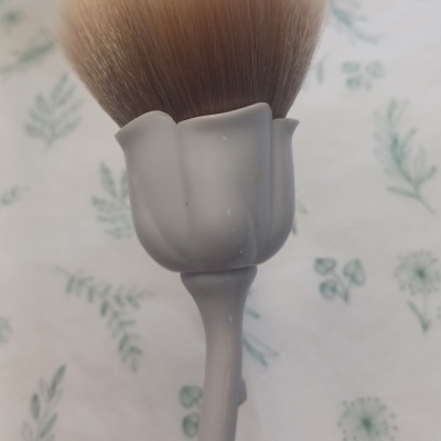 Pincel de maquilhagem com cabo branco formato flor numa superfície com padrão de folhas verde.