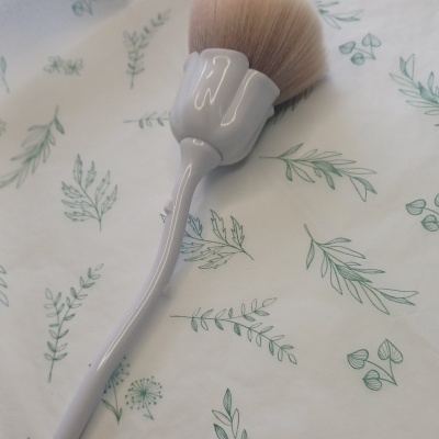 Pincel de maquilhagem com cabo cinza sobre tecido branco com padrão de folhas verdes