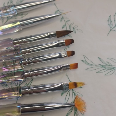 Sete pincéis de unhas com cabo transparente e cerdas variadas sobre papel branco com desenhos de folhas verdes.