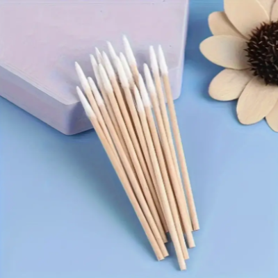 Cotonetes de madeira com pontas de algodão em superfície azul com caixa lilás e flor decorativa.