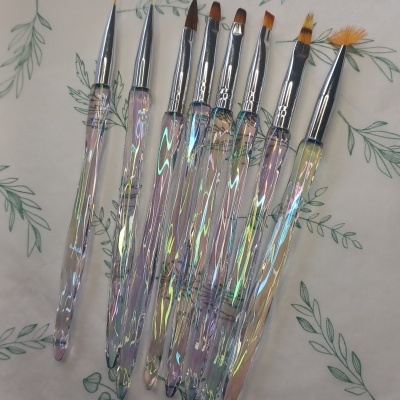 Conjunto de pincéis para unhas com cabos iridescentes e cerdas variadas sobre fundo com padrão de folhas verdes