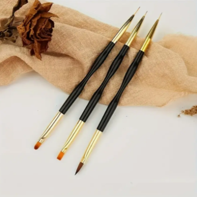 Três pincéis artísticos com cabos pretos e ponteiras douradas sobre tecido bege e flor seca