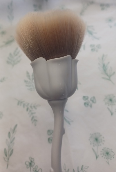 Pincel de maquilhagem com cabo branco formato flor numa superfície com padrão de folhas verde.