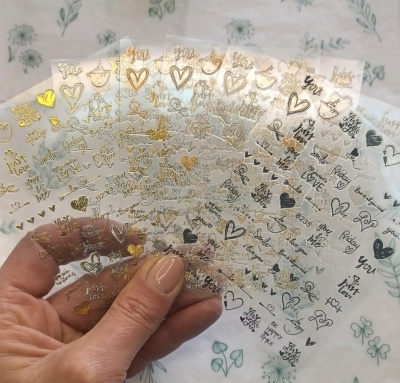 Folhas transparentes com autocolantes metálicos coloridos com corações, estrelas e palavras em inglês.