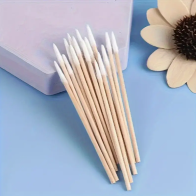 Cotonetes de madeira com pontas de algodão em superfície azul com caixa lilás e flor decorativa.