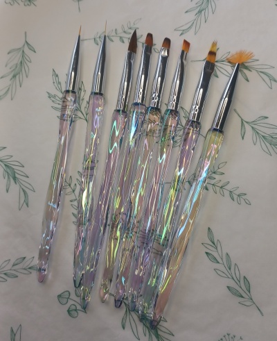 Conjunto de pincéis para unhas com cabos iridescentes e cerdas variadas sobre fundo com padrão de folhas verdes