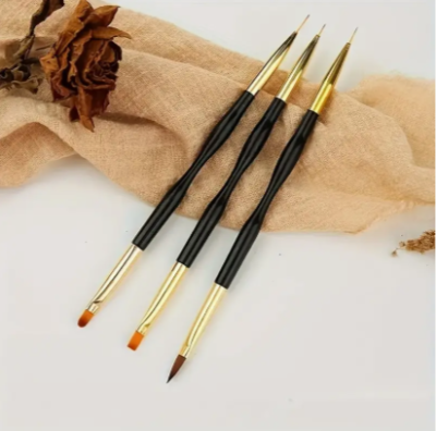 Três pincéis artísticos com cabos pretos e ponteiras douradas sobre tecido bege e flor seca