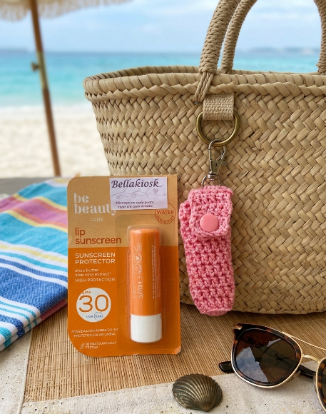 Protetor labial, bolsa de palha com chaveiro rosa, óculos de sol e praia ao fundo