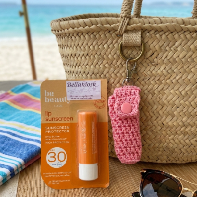 Protetor labial, bolsa de palha com chaveiro rosa, óculos de sol e praia ao fundo
