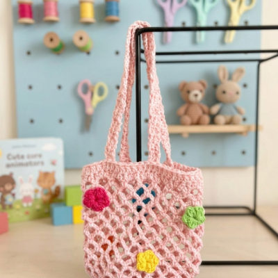 Saco de malha rosa com flores coloridas pendurado em estrutura metálica com fundo de parede azul e objetos de artesanato