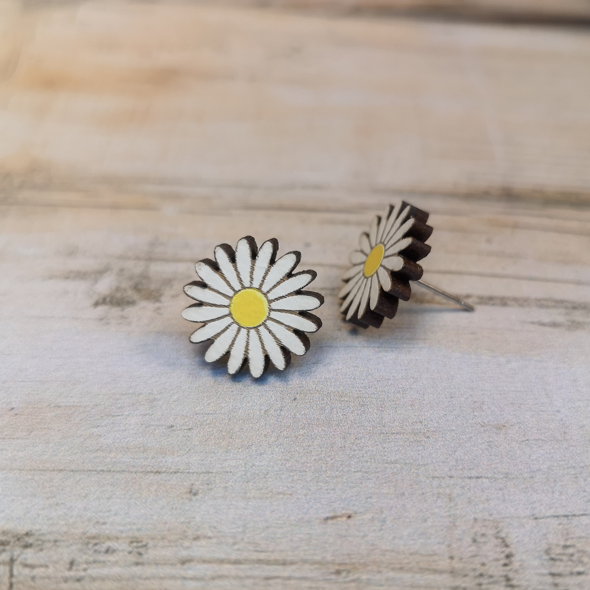 Brincos Margarida Brincos em forma de margaridas brancas com centro amarelo sobre superfície de madeira clara