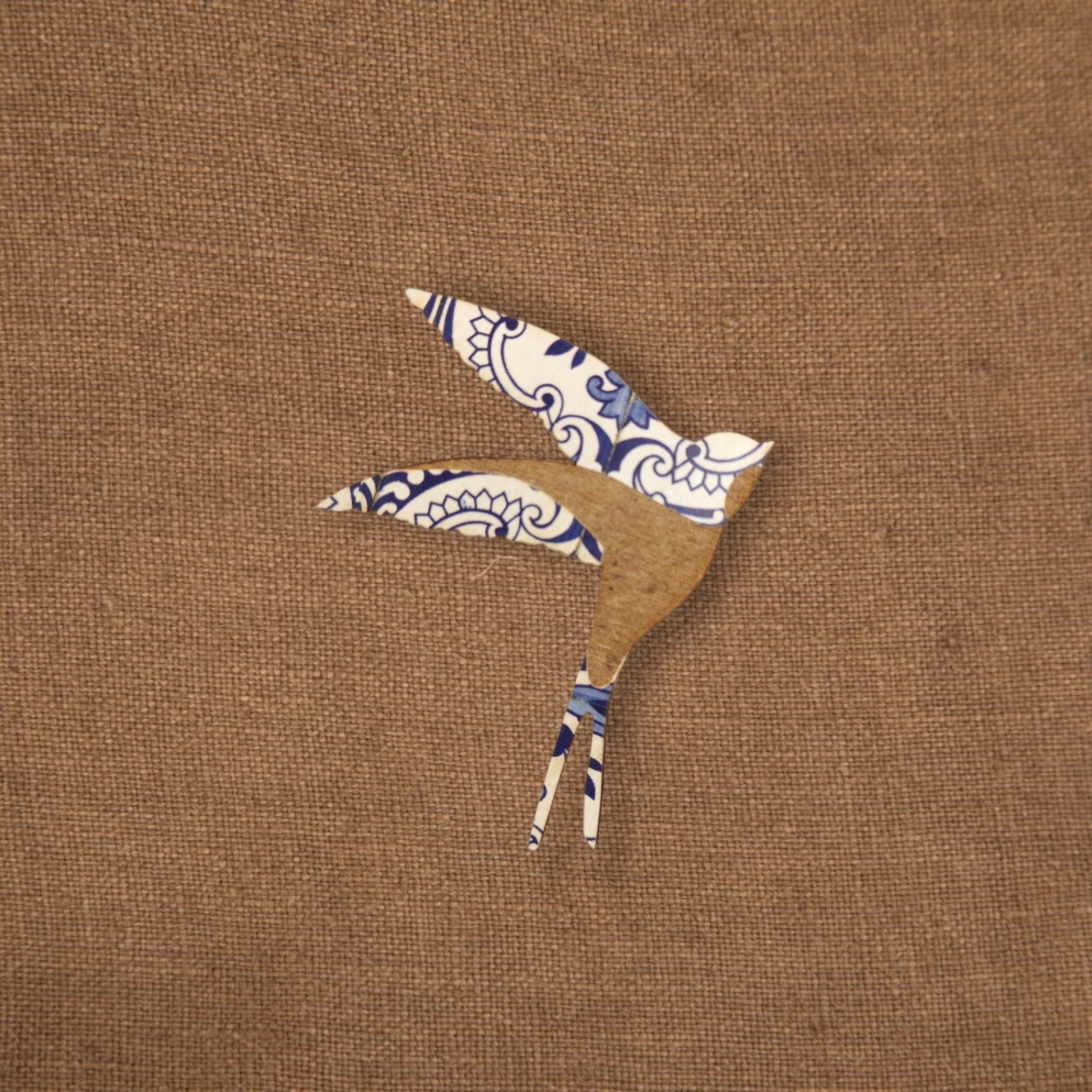 Pregadeira Andorinha Azulejo Recorte em forma de pássaro azul, branco e castanho sobre fundo de tecido bege.