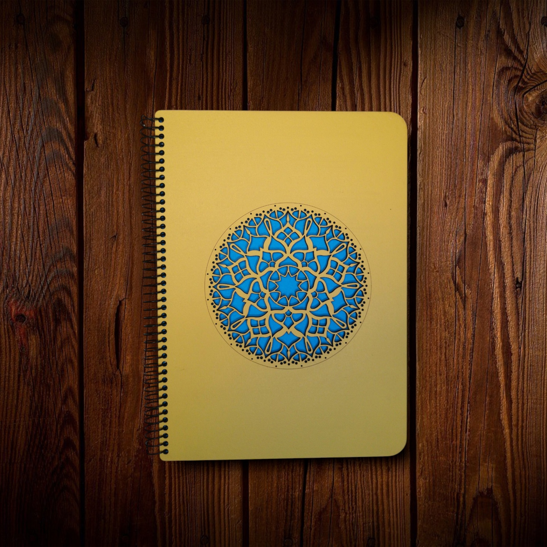 Caderno Mandala Caderno amarelo com padrão circular azul em capa sobre mesa de madeira.