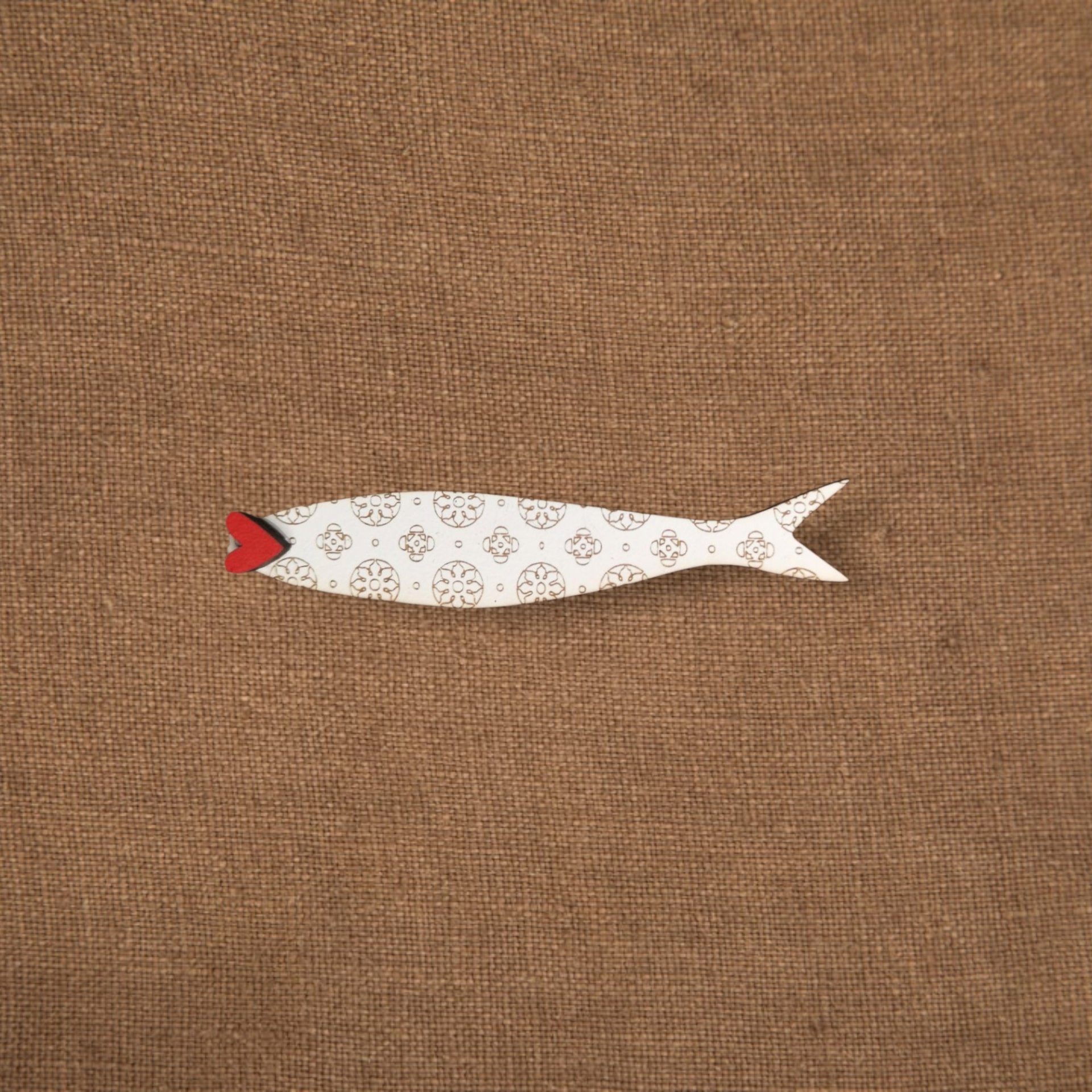 Pregadeira Sardinha Peixe decorativo em madeira branca com padrões castanhos e boca vermelha sobre tecido castanho