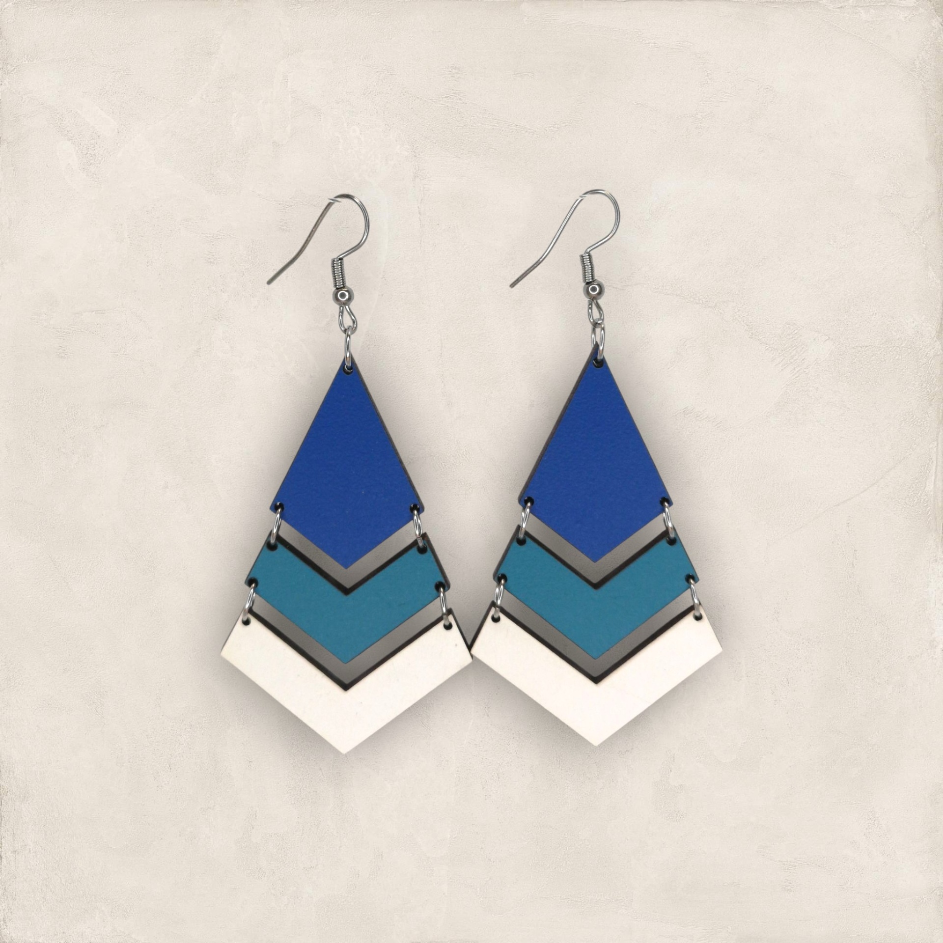 Brincos Rute Brincos pendentes com três lâminas azuis e branca em fundo bege.