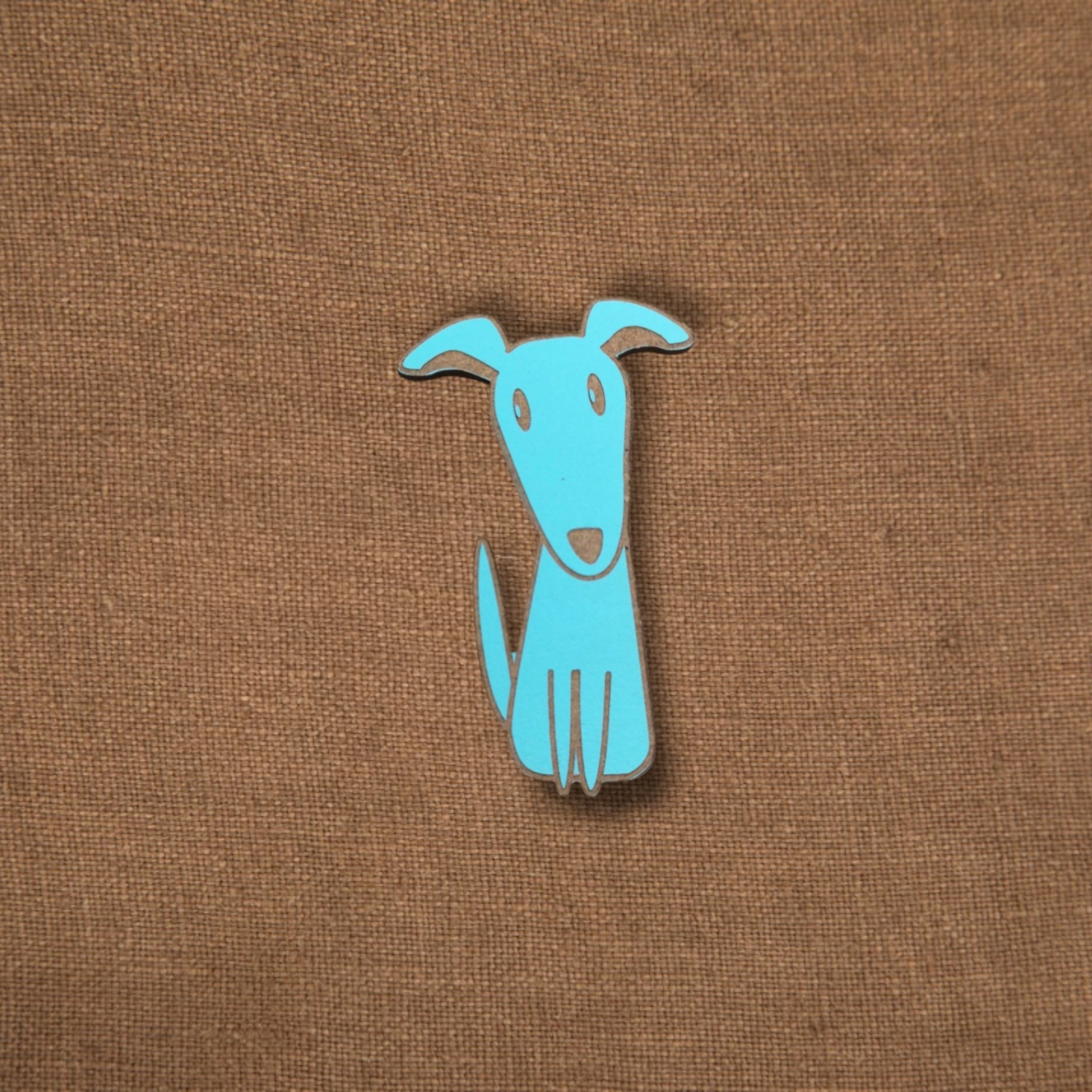 Pregadeira Cão Pin metálico azul em forma de cão estilizado sobre tecido castanho
